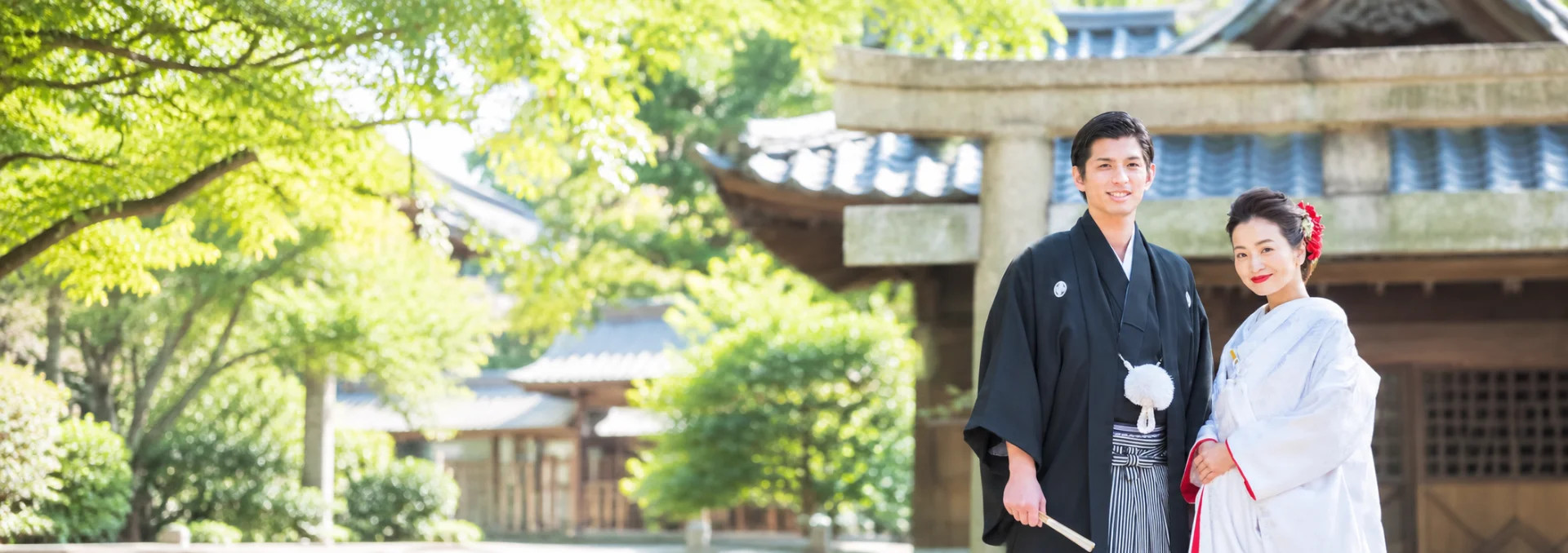新緑に包まれた神社の鳥居の前に並んで立つ、紋付袴と白無垢姿の男女